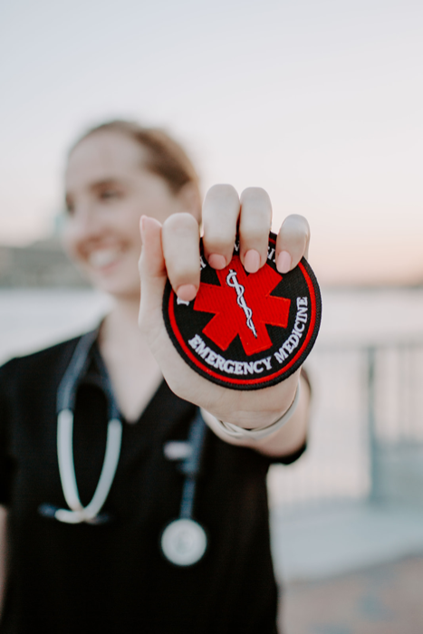 An EMT dressed in black scrubs holds an Emergency Medicine embroidered patch with a medical emblem.