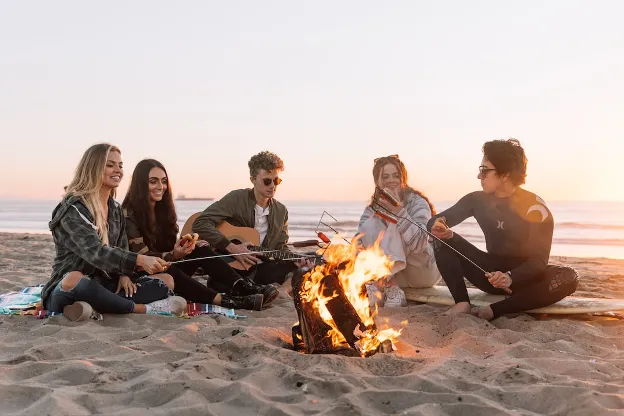 Young adults roasting hotdogs and playing guitar on a beach, representing social connections made during RV life.


