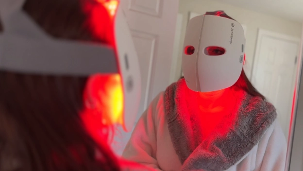 Reflection in a mirror of a woman wearing the white TheraFace mask with red LED lights glowing.