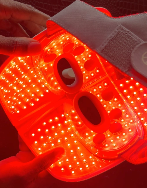 Hands holding the TheraFace mask open to reveal bright red and infrared LED lights illuminating the interior.