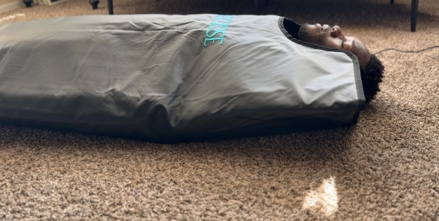 Person relaxing inside HigherDose infrared sauna blanket during heat therapy session on carpet.