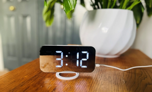 Digital alarm clock on wooden table beside a plant, used as part of a healthy work from home daily routine.