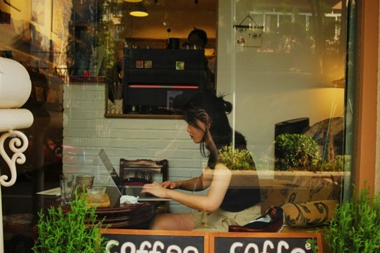 Virtual assistant working on a laptop in a coffee shop showing the risks of public Wi-Fi and unsecured networks.