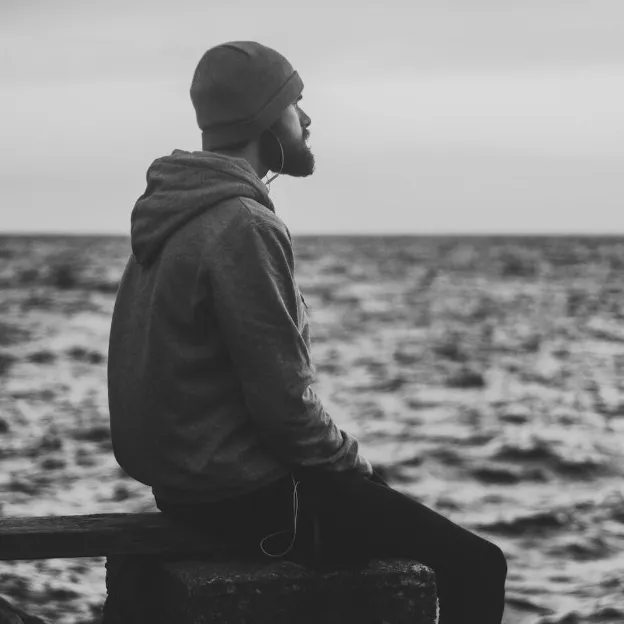 grayscale photo of man in hoodie and kit cap sitting near bodies of water